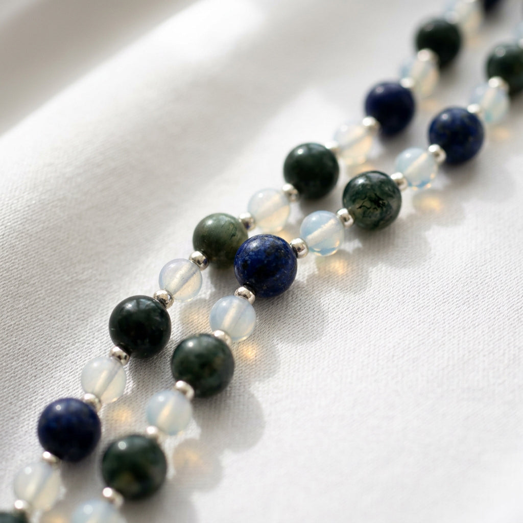 Close-up of two parallel sections of necklace on cream satin, showing dark green peacock pearls, deep blue lapis lazuli and pale translucent moonstone beads separated by small sterling silver spacers.