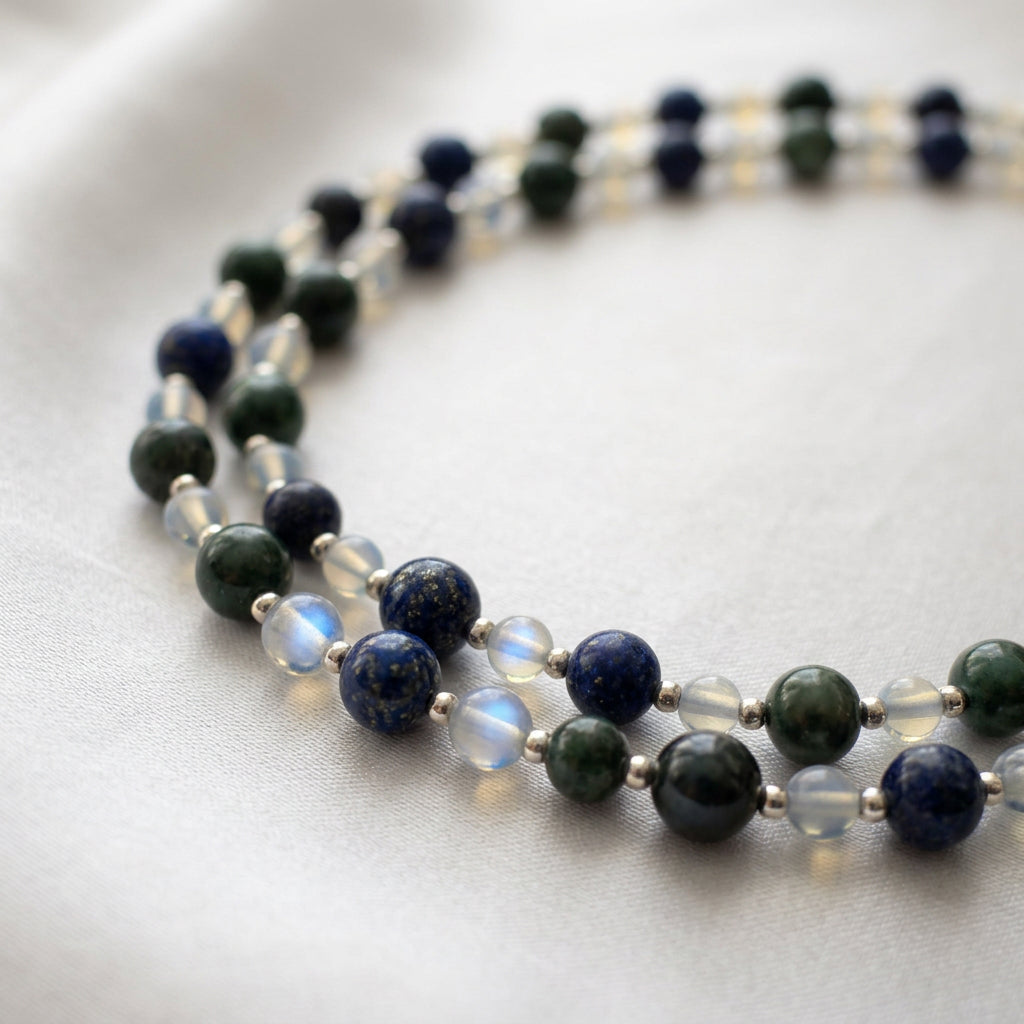 Close-up of loosely coiled dark green peacock pearls, deep blue lapis lazuli and translucent moonstone beads with blue flashes, separated by small sterling silver spacers on cream satin.