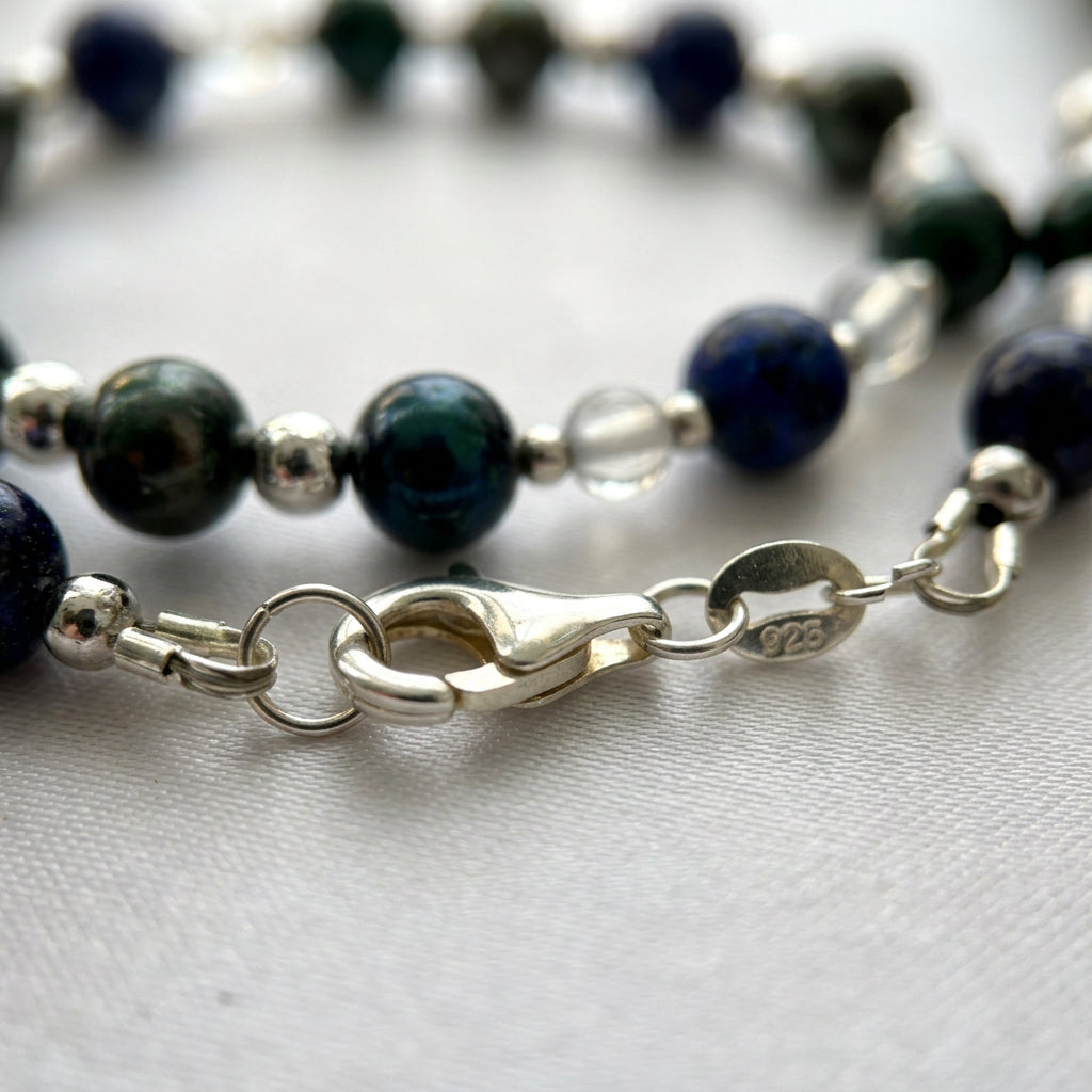 Close-up of a fastened silver lobster clasp and a small tag stamped 925 on cream satin, with dark green peacock pearls, deep blue lapis lazuli, pale moonstone beads and sterling silver spacers behind it.