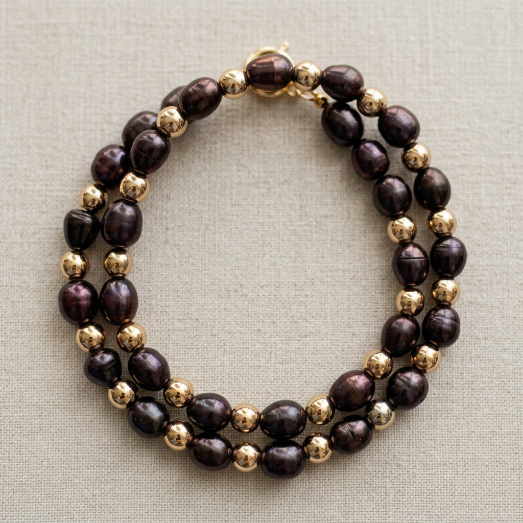 Overhead view of a deep purple rice pearl necklace coiled into two oval loops on cream linen, with irregular lustrous pearls alternating with polished gold-coloured hematite beads.⠋