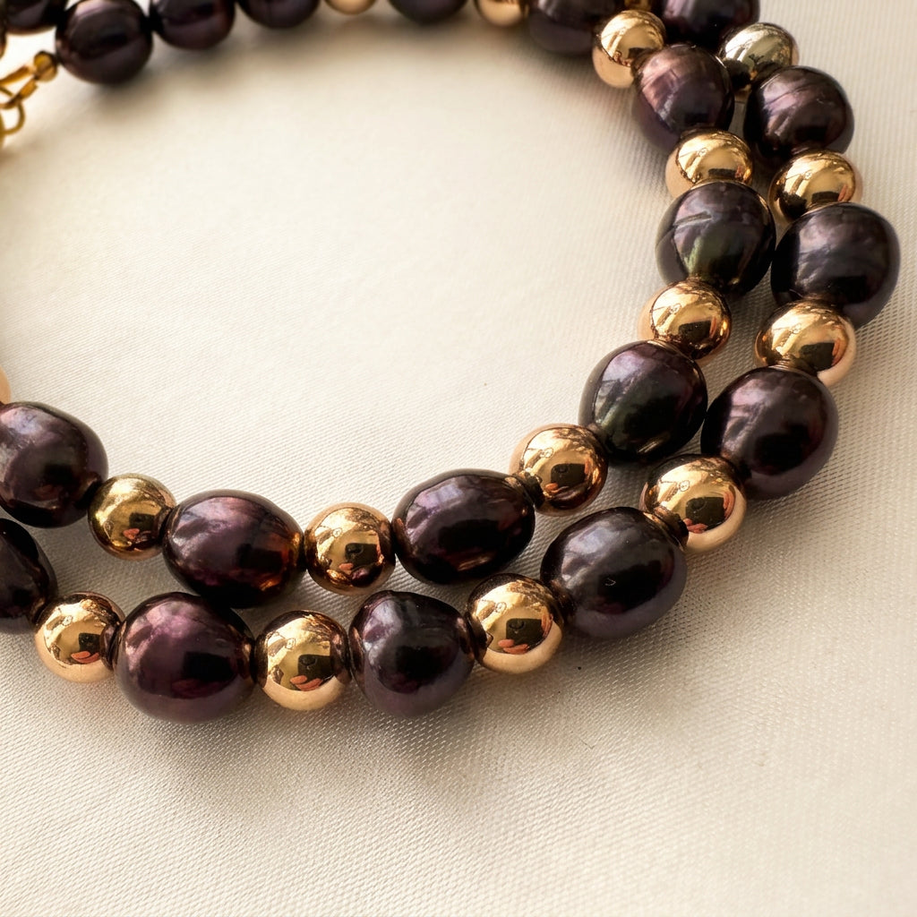 Close-up of a deep purple rice pearl necklace looped on cream satin, highlighting the pearls’ plum-coloured reflections and natural surface ripples beside polished gold-coloured hematite beads.
