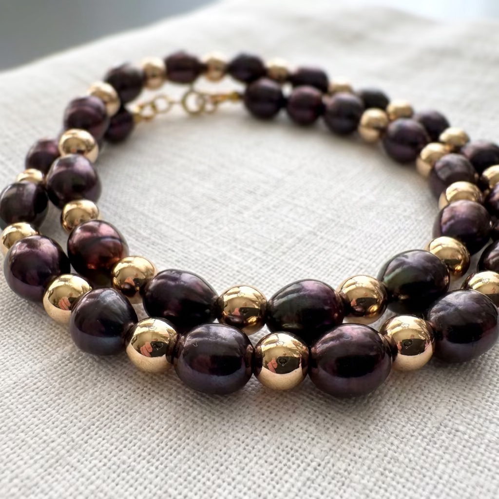 Deep purple rice pearl necklace coiled on cream linen, showing the pearls’ glossy, irregular surfaces alternating with mirror-polished gold-coloured hematite beads.