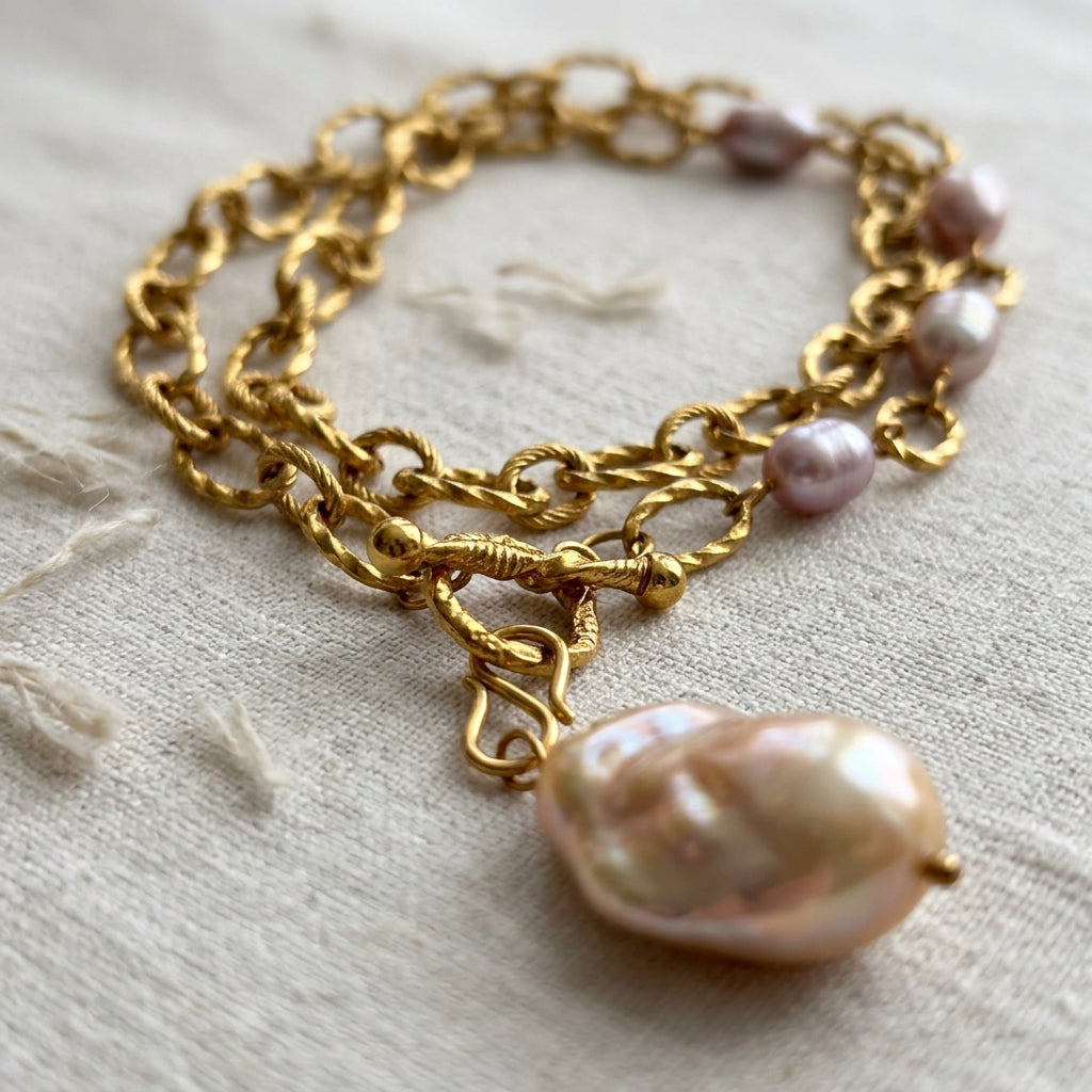 Keshi pearl pendant necklace coiled on cream embroidered linen, with a large lustrous cream pearl, textured gold-coated oval links, four mauve-white rice pearls and a fastened toggle clasp.