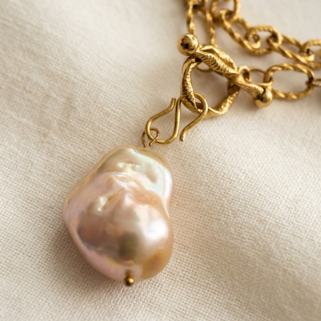 Close-up of a large cream keshi pearl pendant with raised nacre folds and pink-gold reflections, suspended below a fastened textured gold-coated toggle clasp on cream linen.