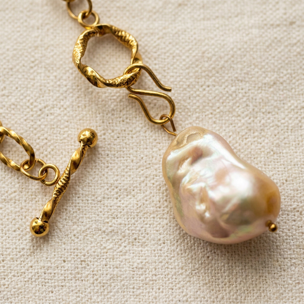 Close-up of an unfastened textured gold-coated toggle clasp, with the separate bar beside the ring and a large cream keshi pearl pendant showing pink-green iridescence on cream linen.