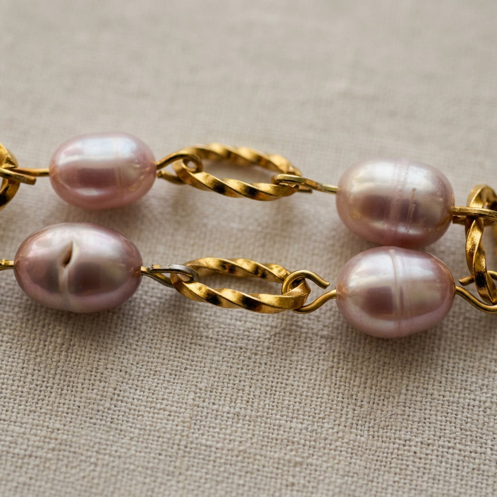 Close-up of four lustrous mauve-pink freshwater rice pearls connected by textured gold-coated oval links on cream linen, showing their natural surface ridges and variations.