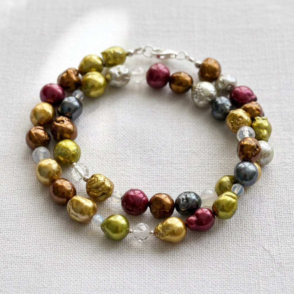 Multicoloured baroque pearl necklace loosely coiled on pale linen, showing lime-gold, bronze, burgundy and silver-grey pearls, pale translucent gemstone beads and a silver clasp near the top.