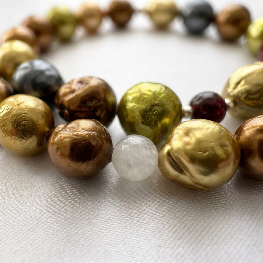 Close-up of bronze, gold and lime-green baroque pearls with rippled surfaces beside a milky moonstone bead, with a dark red garnet and silver-grey pearl behind them on pale satin.