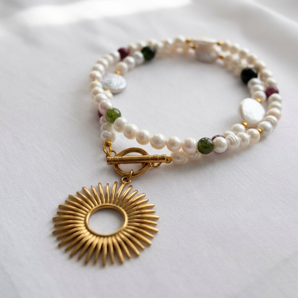 White baroque and coin pearl necklace loosely coiled on cream fabric, with red and green tourmaline accents, a fastened gold-plated toggle clasp and an open-circle sunburst pendant in the foreground.⠋