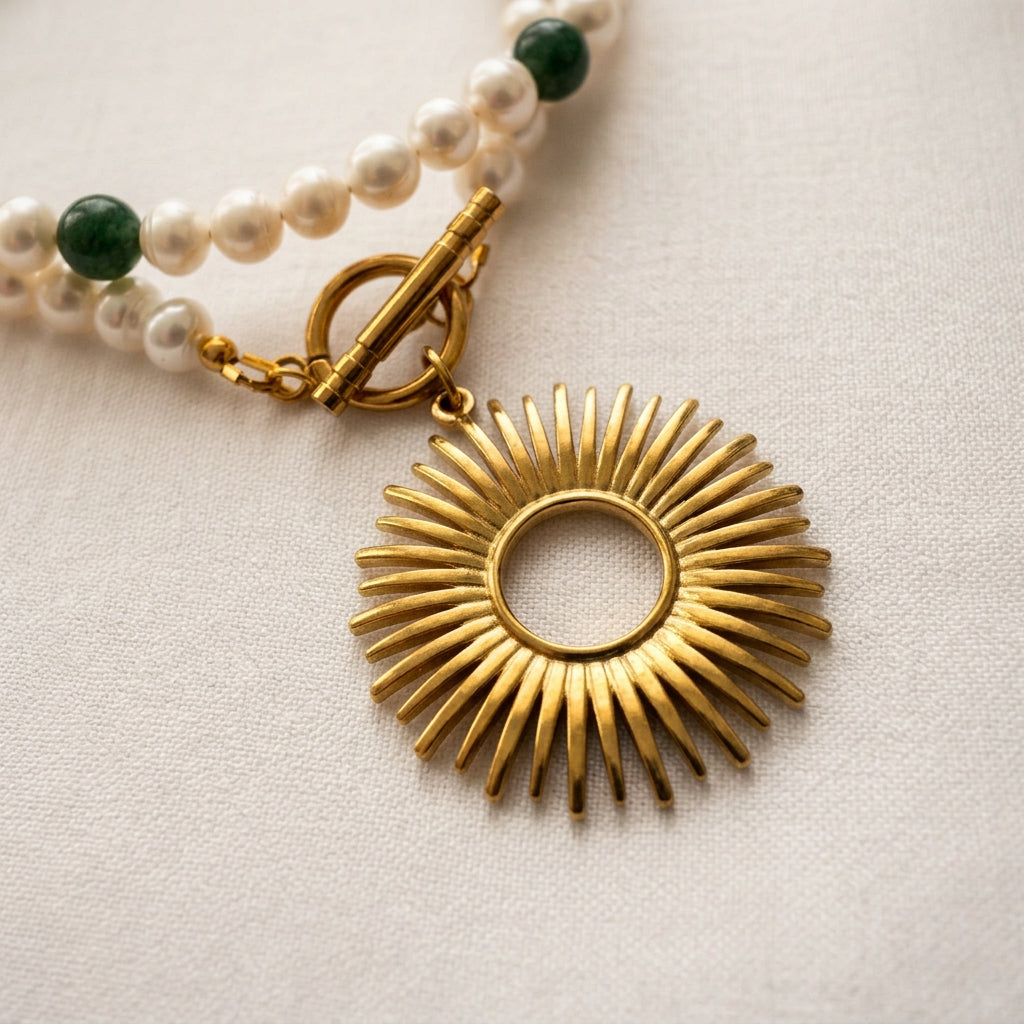 Close-up of an open-circle sunburst pendant and fastened toggle clasp in gold-plated stainless steel, with white baroque pearls and green tourmaline beads on cream fabric.