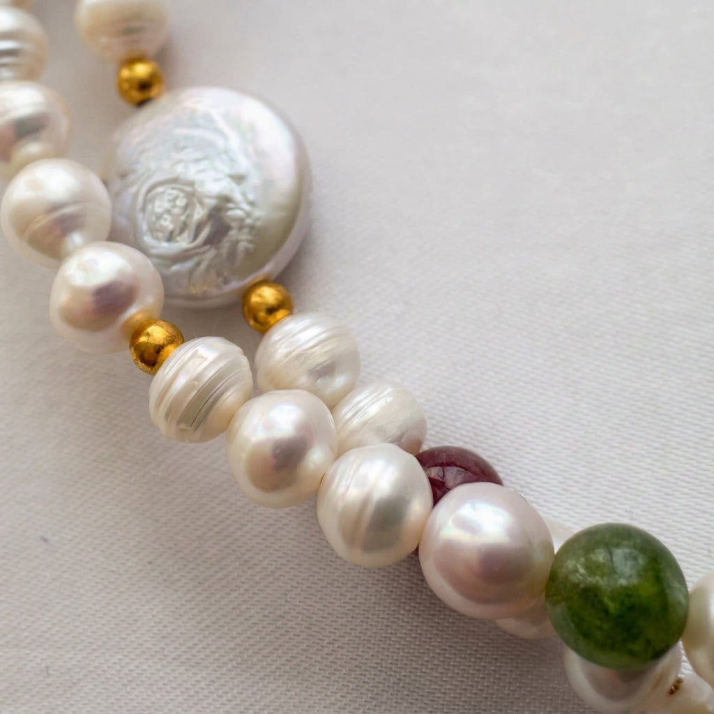 Close-up of lustrous white baroque pearls, a textured flat coin pearl and red and green tourmaline beads, with gold-coloured spacer accents on cream fabric.