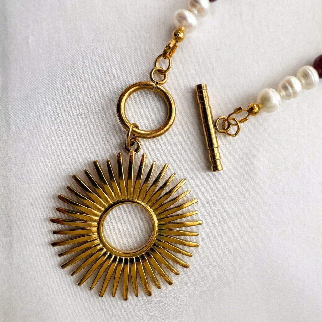 Close-up of a gold-plated stainless steel sunburst pendant and unfastened toggle clasp, with the ring and bar separated beside white baroque pearls on cream fabric.⠋