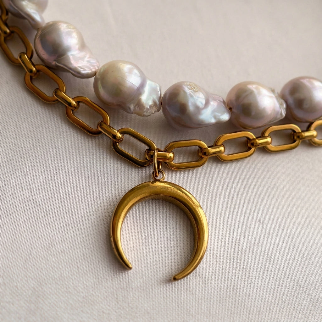 Close-up of a downward-pointing gold-plated crescent moon pendant on a chunky paperclip chain, with large irregular silvery-white keshi pearls above it on cream satin.