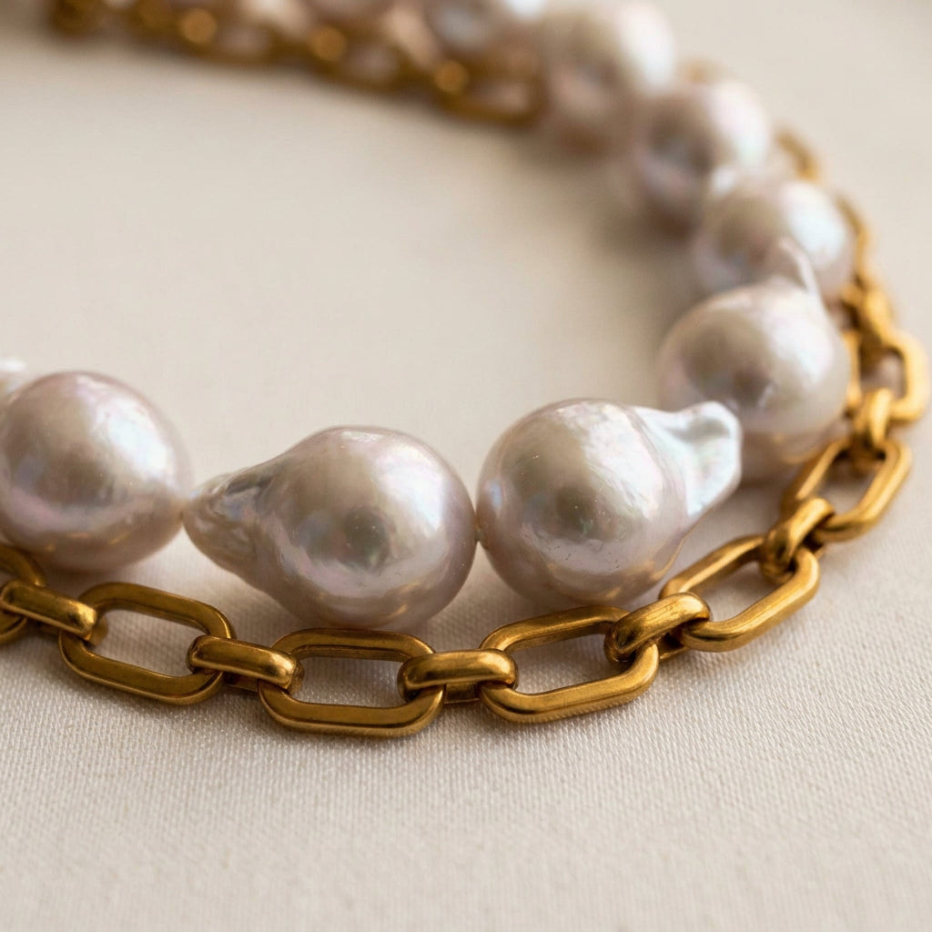 Close-up of large silvery-white keshi pearls with irregular petal-like edges beside a chunky gold-plated paperclip chain, arranged in parallel curves on cream satin.