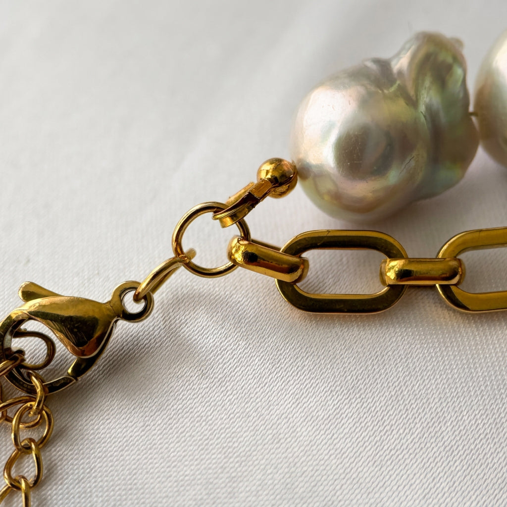 Close-up of a fastened gold-plated lobster clasp and connecting ring joining a chunky paperclip chain and silvery-white keshi pearl strand on cream satin.