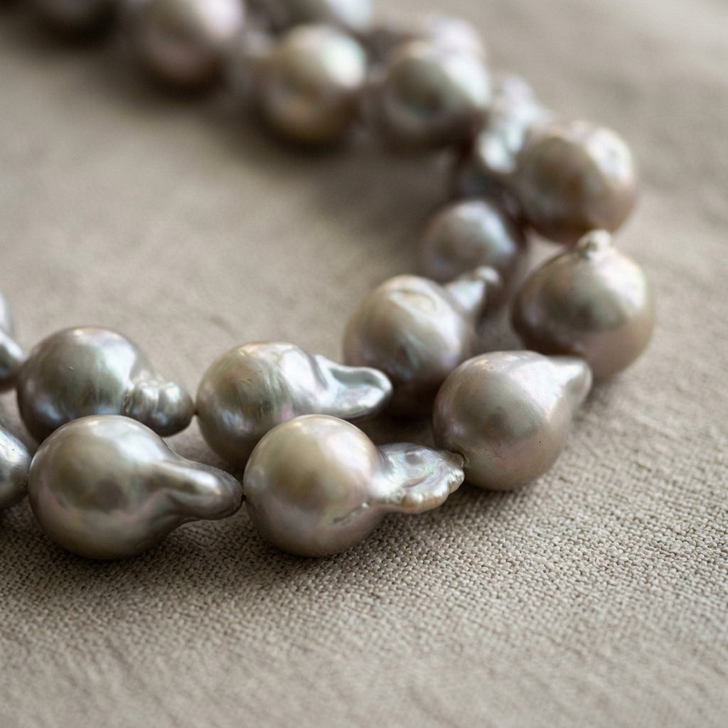 Close-up of loosely coiled silvery-white keshi pearls on grey linen, showing rounded centres, irregular petal-like edges and soft green and pink iridescence.