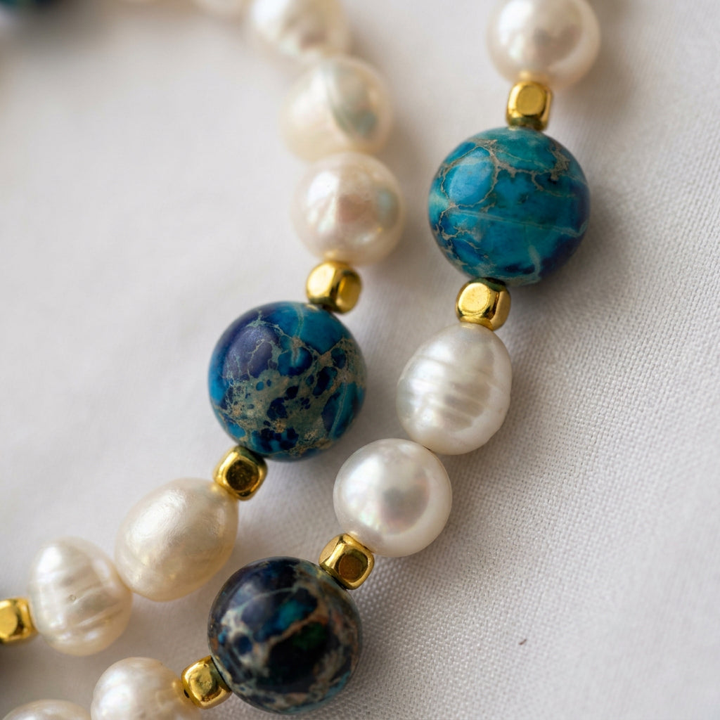 Blue jasper and white baroque pearl necklace loosely coiled on cream satin, with gold-coloured hematite spacers, a fastened toggle clasp and a large sculpted gold-plated flower pendant in the foreground.