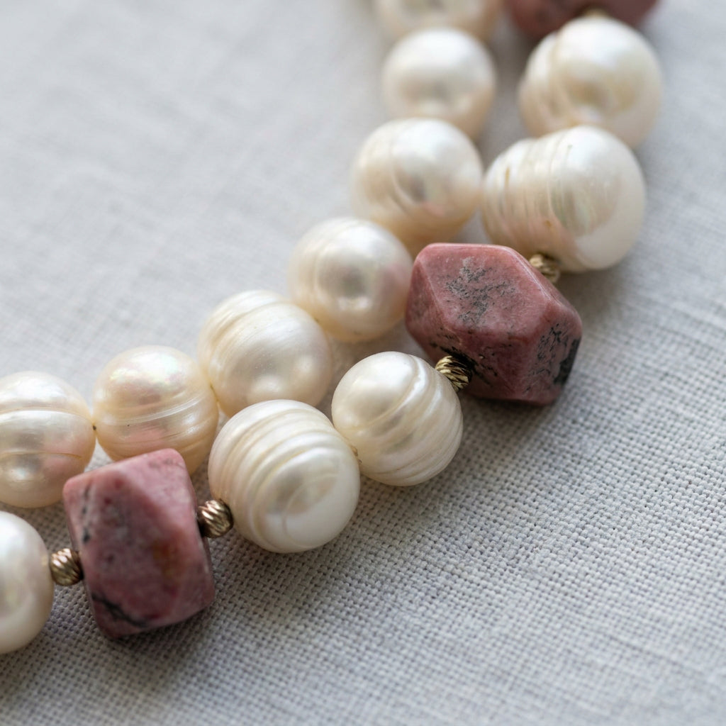 Close-up of creamy white baroque pearls with pronounced natural ridges beside irregular pink opal beads with dark markings, separated by textured sterling silver spacers on grey linen.⠋