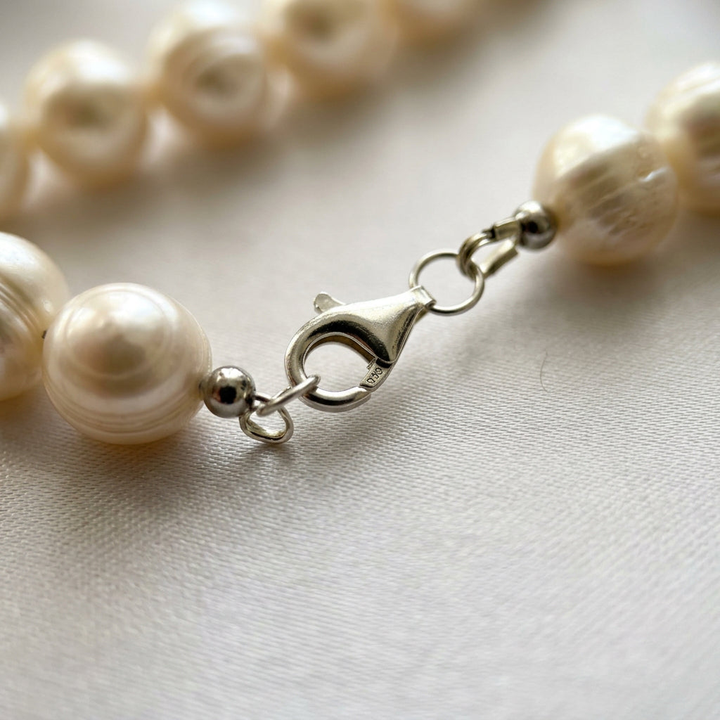 Close-up of a fastened sterling silver lobster clasp stamped 925, joining large creamy white baroque pearls on pale satin.