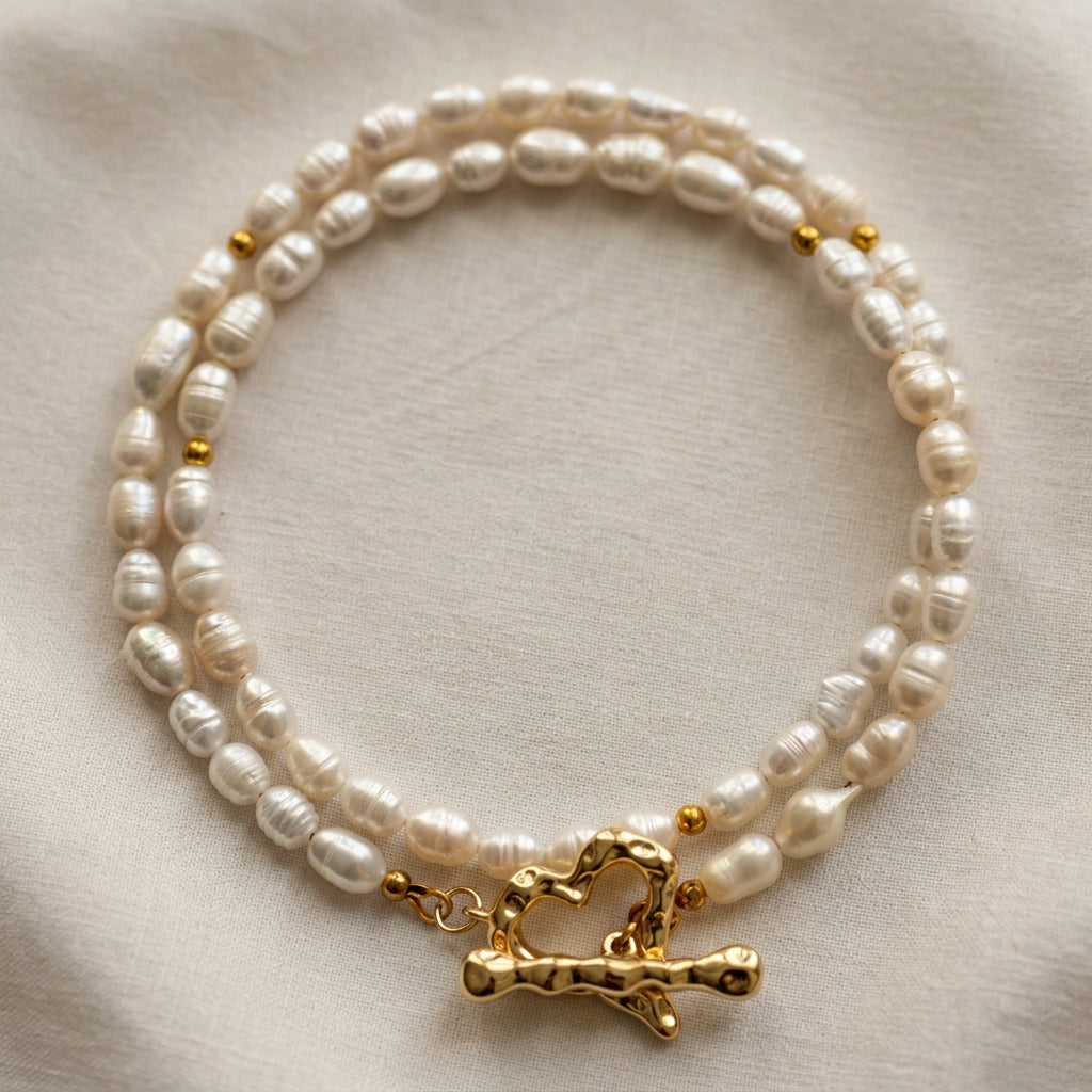 White rice-shaped baroque pearl necklace loosely coiled on cream fabric, with small gold-plated spacer beads and a fastened, rippled heart-shaped toggle clasp in the foreground.