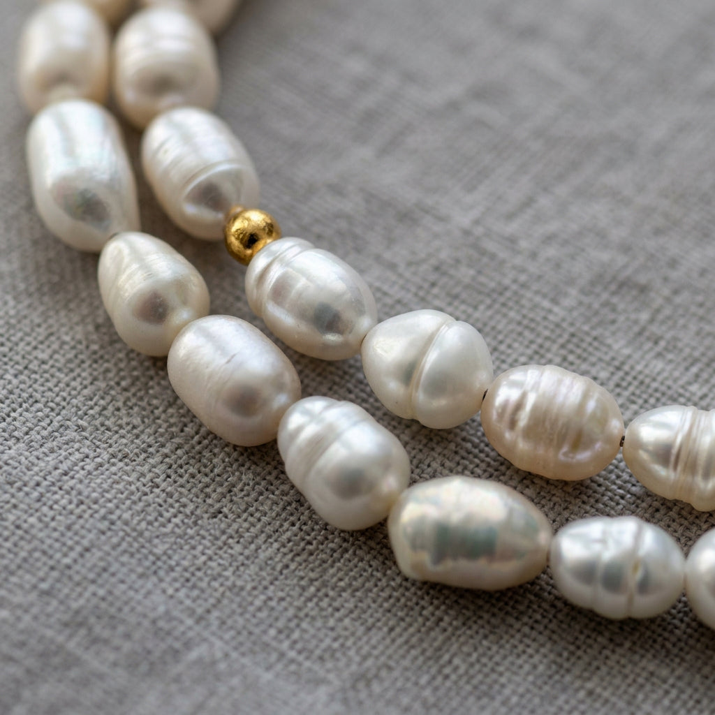 Close-up of a fastened gold-plated stainless steel heart-shaped toggle clasp with a rippled, polished surface, framed by white baroque pearls and small gold-plated beads on cream fabric.