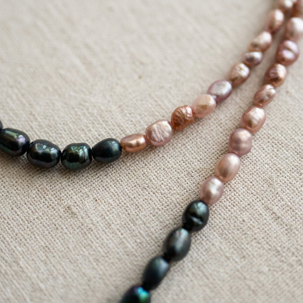 Close-up of blush-pink and dark blue rice pearls on cream linen, showing the transition between colour sections and the pearls’ irregular shapes, ridged surfaces and green-blue lustre.