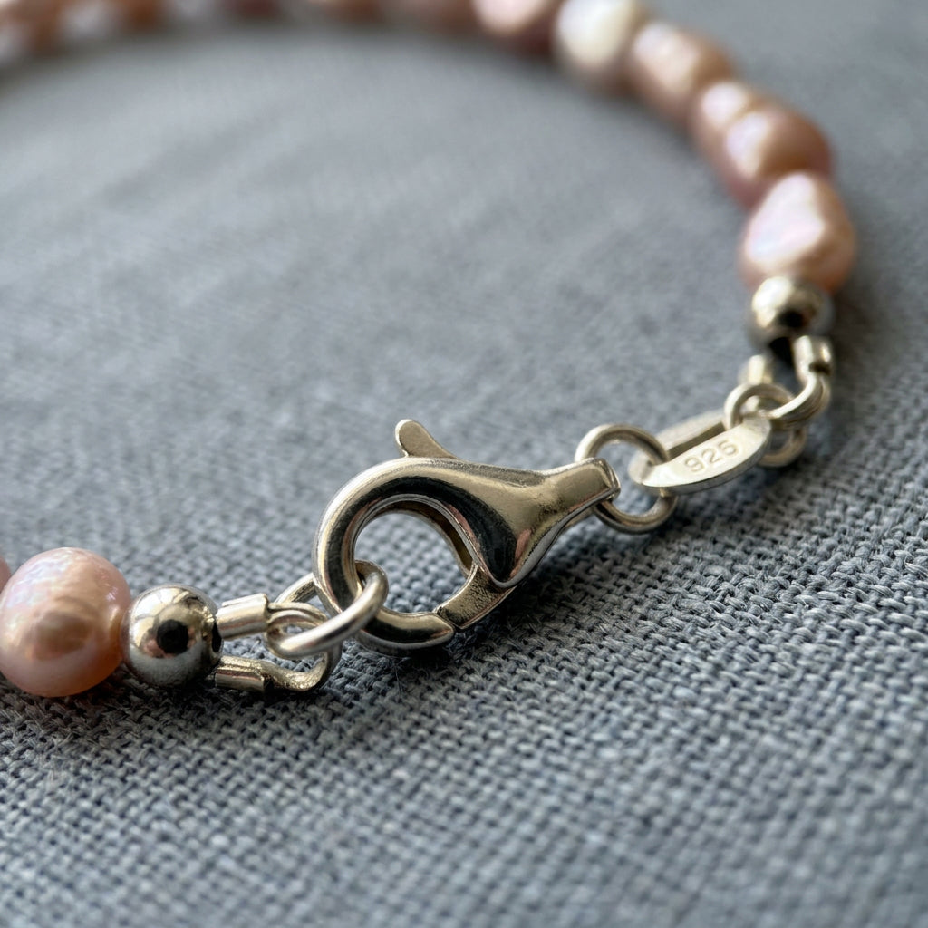 Close-up of a fastened sterling silver lobster clasp and a small tag stamped 925, joining blush-pink rice pearls on textured grey linen.