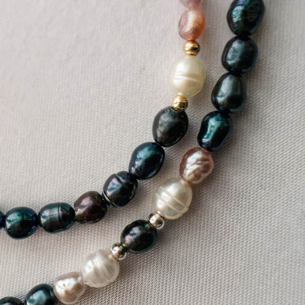 Close-up of dark blue, creamy white and blush-pink rice pearl strands on cream fabric, showing green-blue reflections, natural surface ridges and small metallic spacer accents.