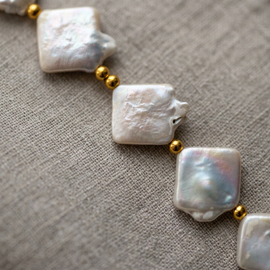 Close-up of rectangular white baroque pearls separated by small gold-plated spacer beads on cream linen, showing their irregular edges, natural surface textures and pink-silver iridescence.