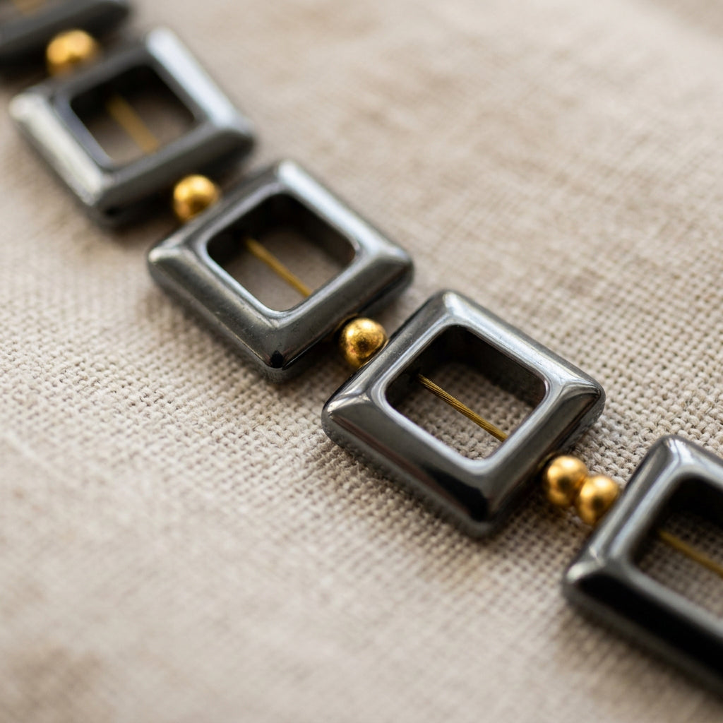 Close-up of glossy black open-square hematite beads separated by small gold-plated spacer beads on cream linen, with the threading visible through each square centre.