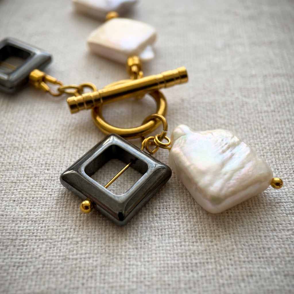 Close-up of a black open-square hematite pendant and iridescent white rectangular baroque pearl pendant beneath a fastened gold-plated toggle clasp on cream linen.