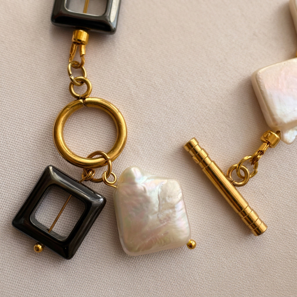 Close-up of an unfastened gold-plated toggle clasp, showing the separate bar and circular ring beside a white rectangular baroque pearl pendant and black open-square hematite pendant on cream fabric.