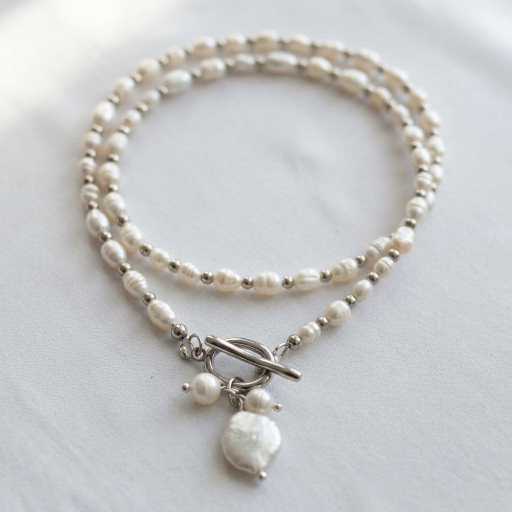 White rice pearl necklace with silver-coloured spacer beads arranged in overlapping loops on cream fabric, with a fastened stainless steel toggle clasp and three-pearl pendant cluster featuring a large coin pearl.