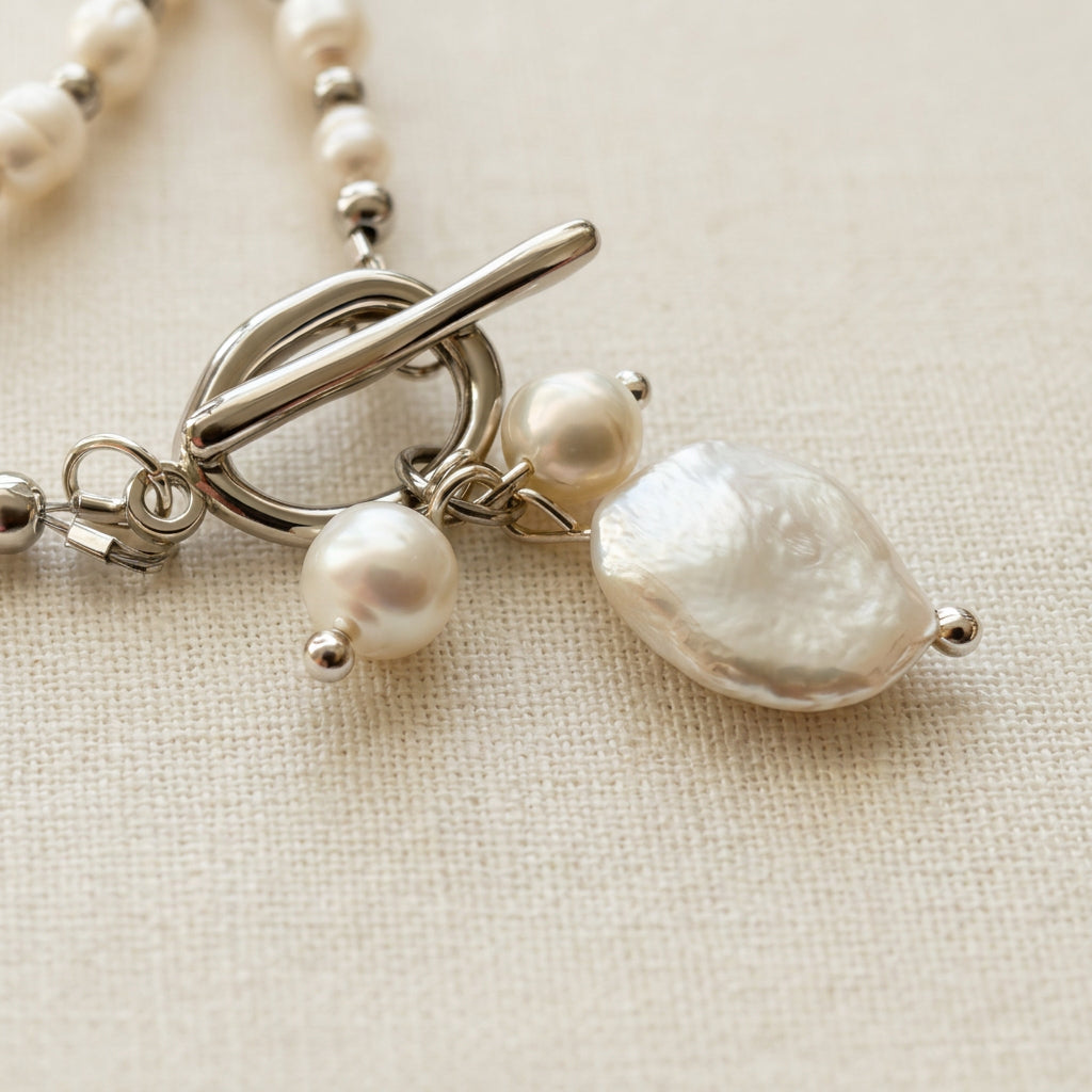Close-up of a fastened stainless steel toggle clasp with three dangling white pearls, including a large textured coin pearl and two smaller baroque pearls, on cream linen.
