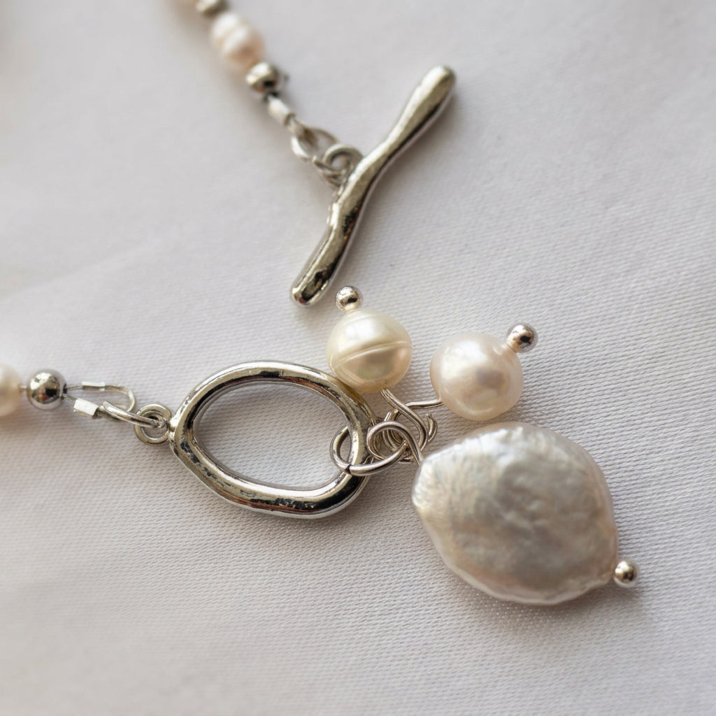 Close-up of an unfastened stainless steel toggle clasp, showing the separate bar and ring beside a pendant cluster of one large white coin pearl and two smaller baroque pearls on cream fabric.