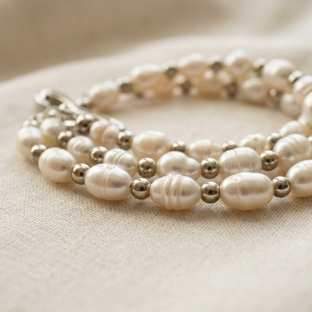 Close-up of white rice pearls alternating with polished silver-coloured spacer beads, arranged in overlapping curves on cream linen, showing the pearls’ natural ridges and lustre.
