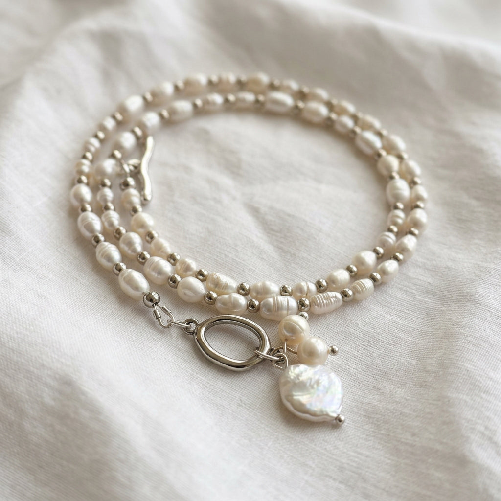 White rice pearl necklace with silver-coloured spacer beads coiled on cream linen, showing an unfastened toggle clasp and a three-pearl pendant cluster with a large white coin pearl.⠋