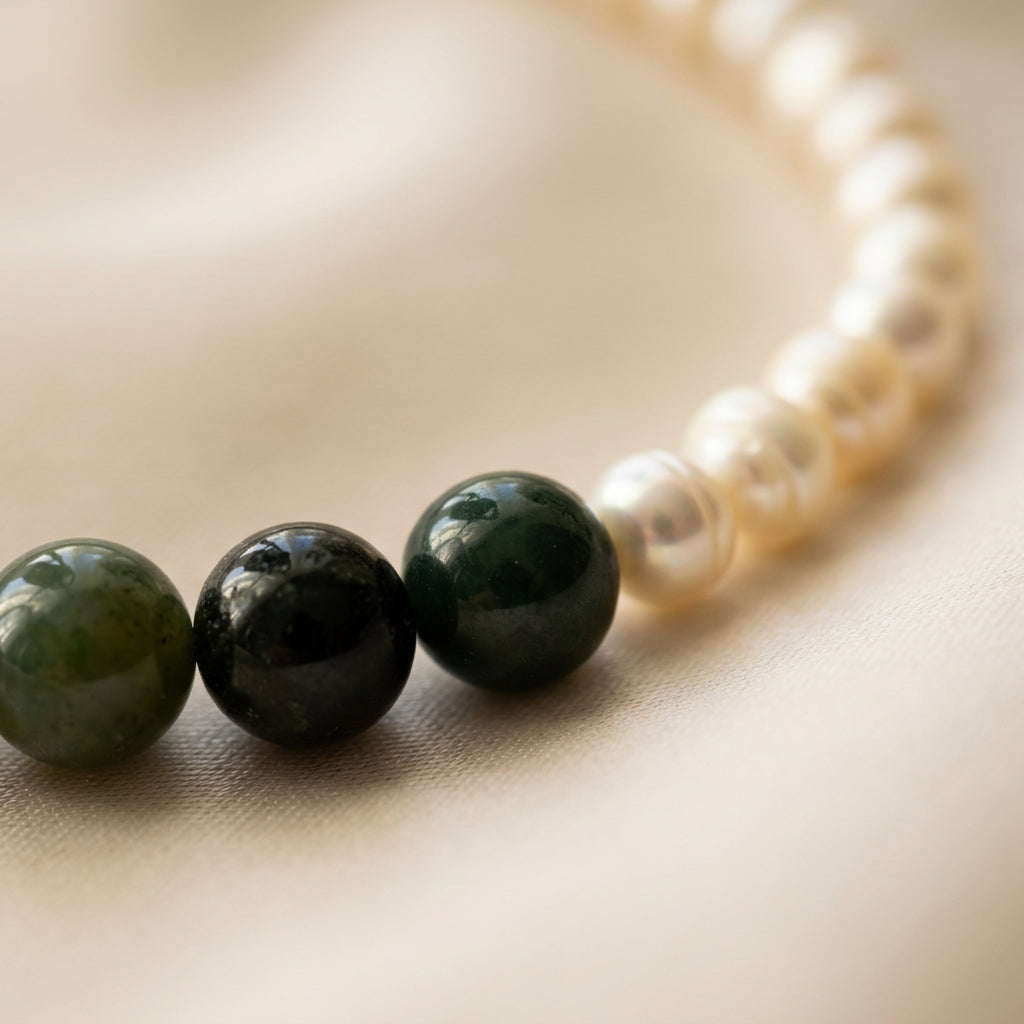 Close-up of polished green obsidian beads transitioning into lustrous white baroque pearls, arranged in a curve on cream fabric.