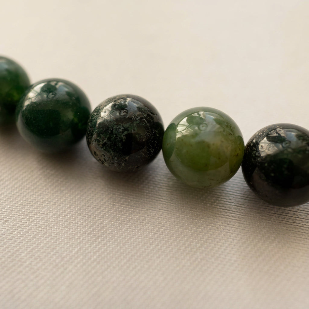 Close-up of polished round green obsidian beads on cream fabric, showing varied shades from olive to deep forest green and naturally mottled surfaces.