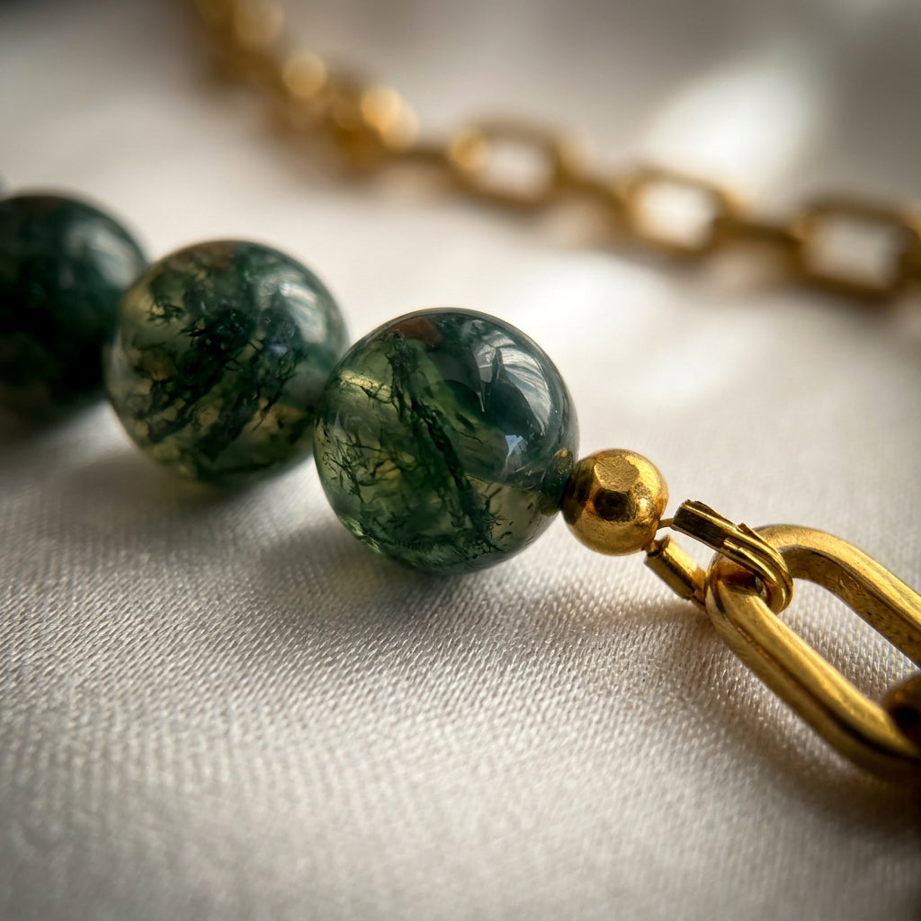 Close-up of translucent green obsidian beads with dark branching patterns, joined to a gold-plated paperclip chain beside a small gold-coloured spacer bead on cream fabric.