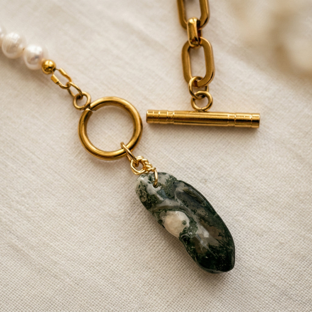 Close-up of an unfastened gold-plated toggle clasp with its separate bar and ring, beside an elongated green Indian agate pendant with cream inclusions on cream linen.