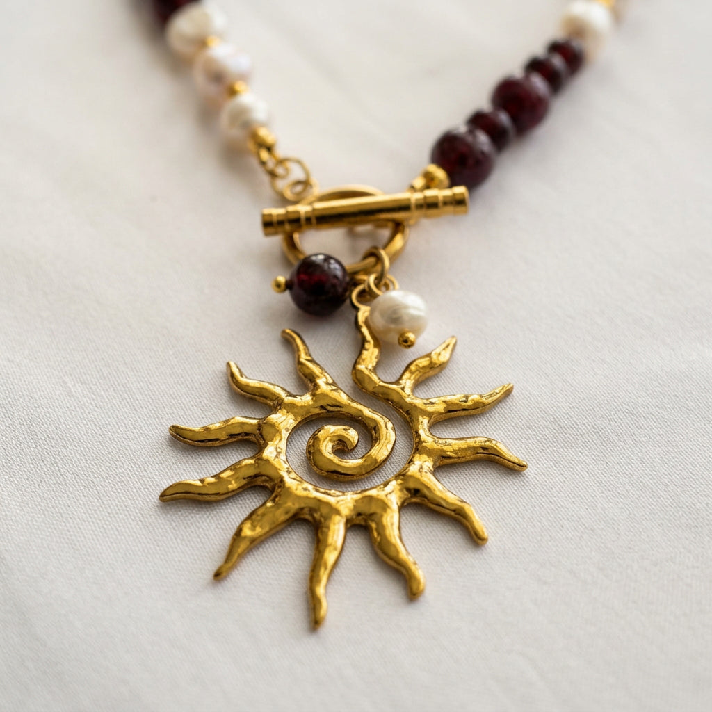 Close-up of a textured gold-coloured sun pendant with a spiral centre and irregular rays, beneath a fastened gold-plated toggle clasp with garnet and white pearl drops on cream fabric.⠋