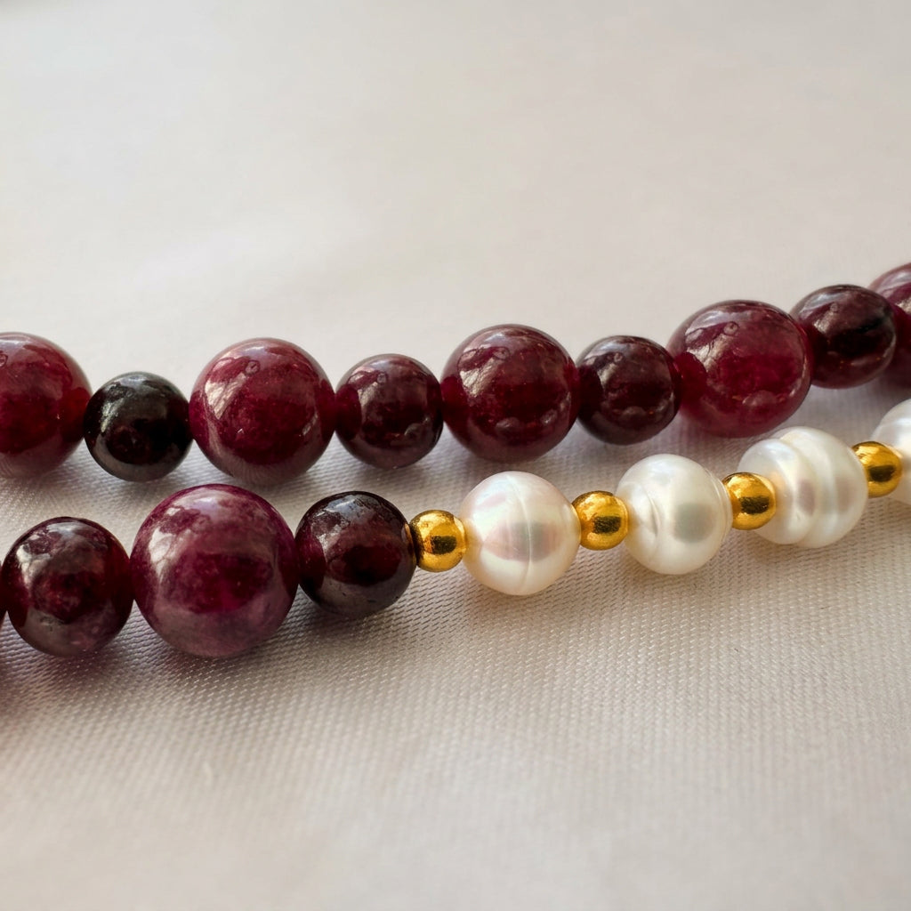 Close-up of alternating large and small burgundy garnet beads beside white baroque pearls separated by gold-plated spacer beads, arranged on cream fabric.