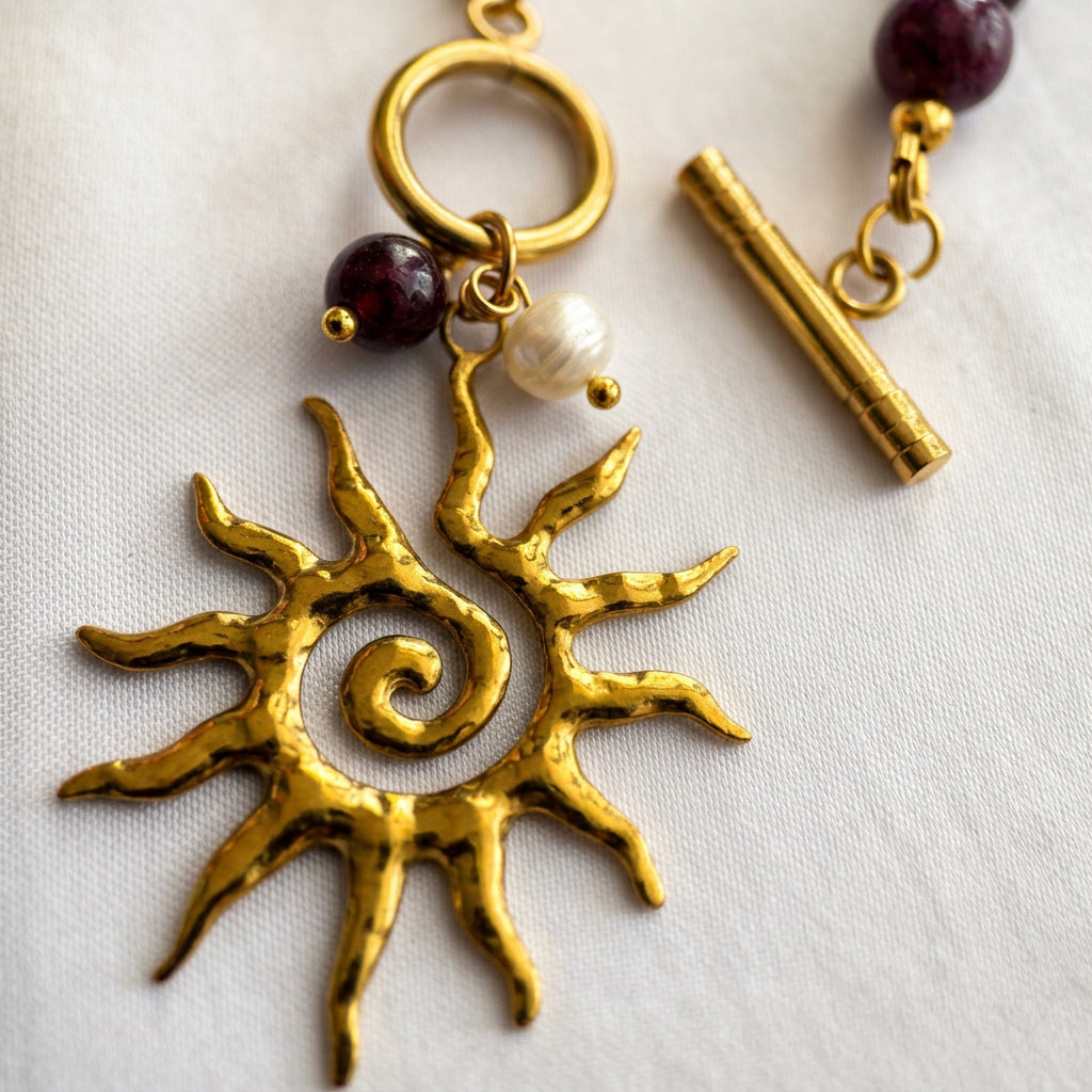 Close-up of a textured gold-coloured spiral sun pendant with garnet and white pearl drops, beside an unfastened gold-plated toggle clasp showing its separate bar and ring on cream fabric.