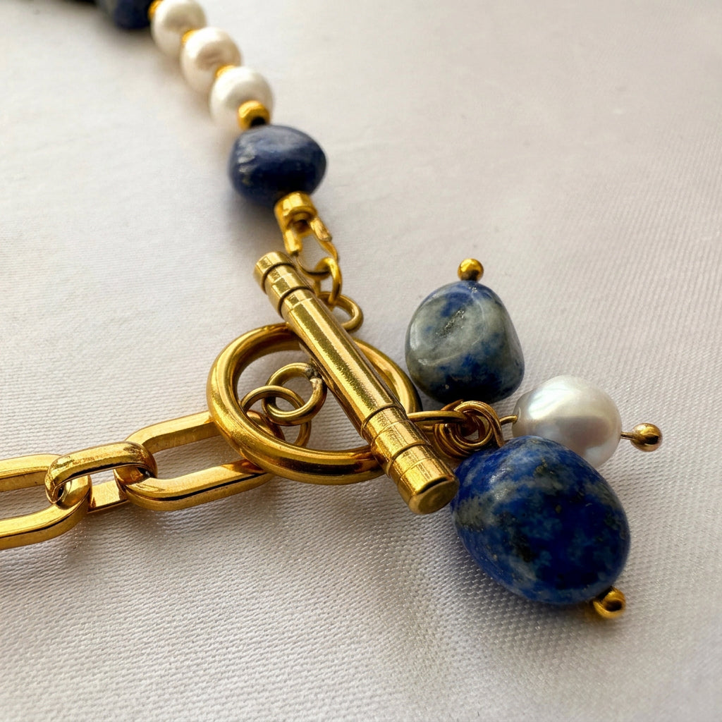 Close-up of a fastened gold-plated toggle clasp beside two blue lapis lazuli drops and one white baroque pearl, with paperclip chain and pearl beads resting on cream fabric.