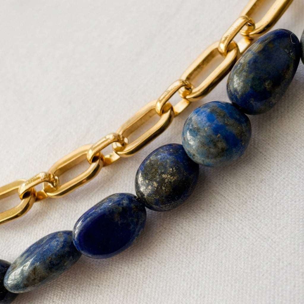Close-up of irregular blue lapis lazuli stones with grey mottling and metallic flecks, arranged beside a gold-plated paperclip chain on cream fabric.