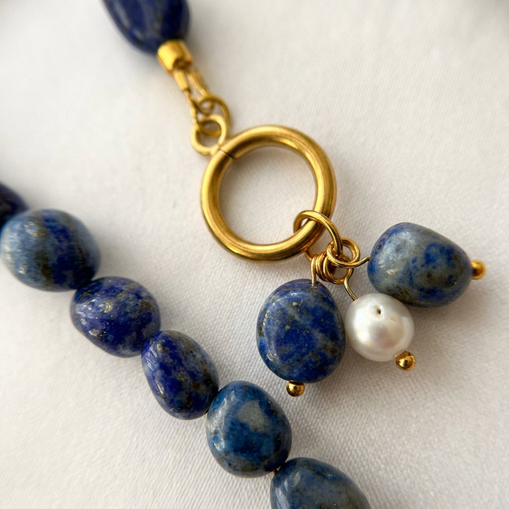 Close-up of two mottled blue lapis lazuli drops and one white baroque pearl hanging from a gold-plated toggle ring, with matching lapis beads on cream fabric.⠋