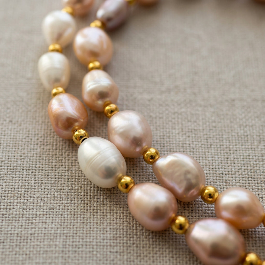 Close-up of white, blush pink and lilac baroque pearls separated by small gold-plated spacer beads on grey linen, showing their irregular shapes, natural ridges and soft lustre.
