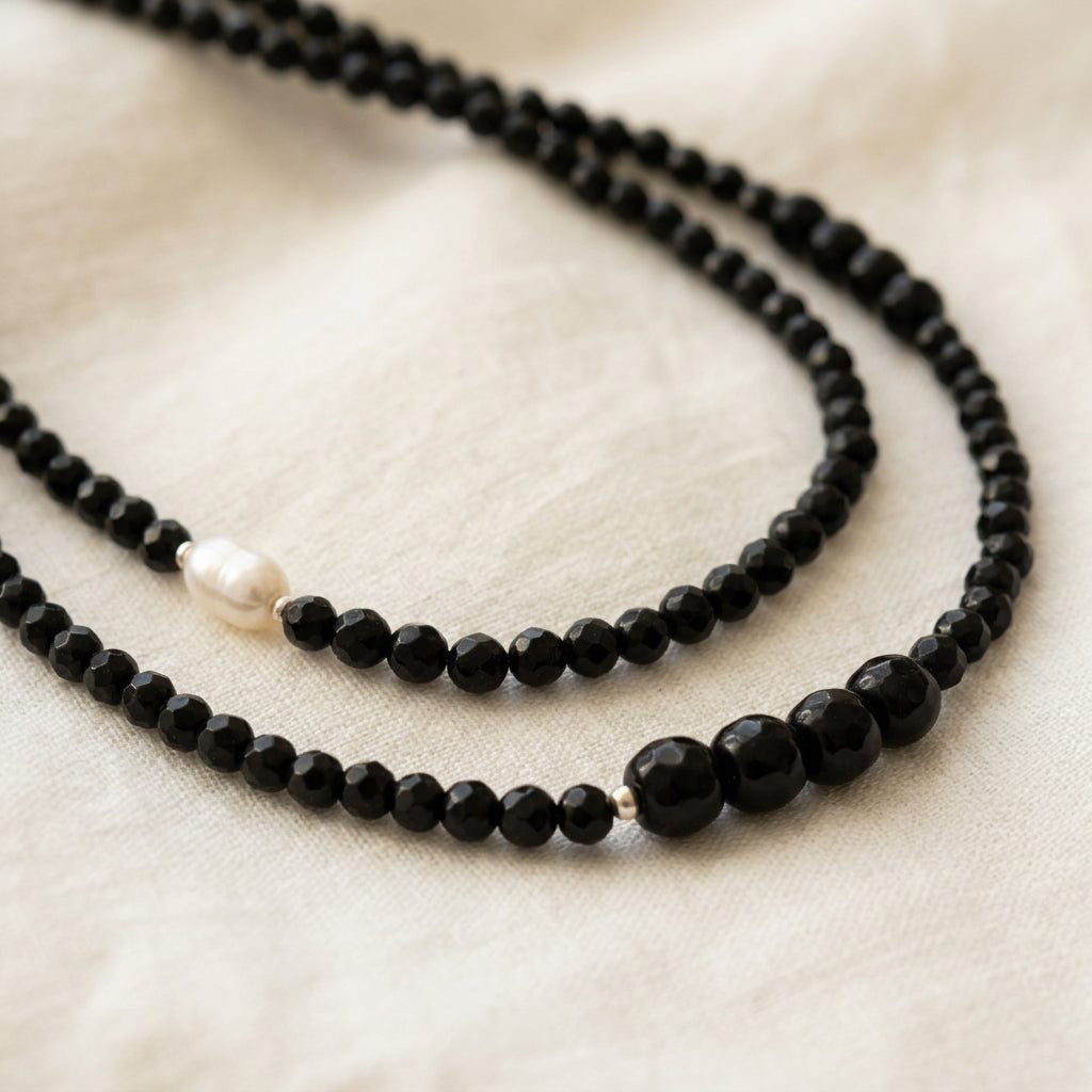 Close-up of two faceted black onyx necklaces on cream fabric, showing a white baroque pearl on one strand and larger black beads with silver spacers on the other.