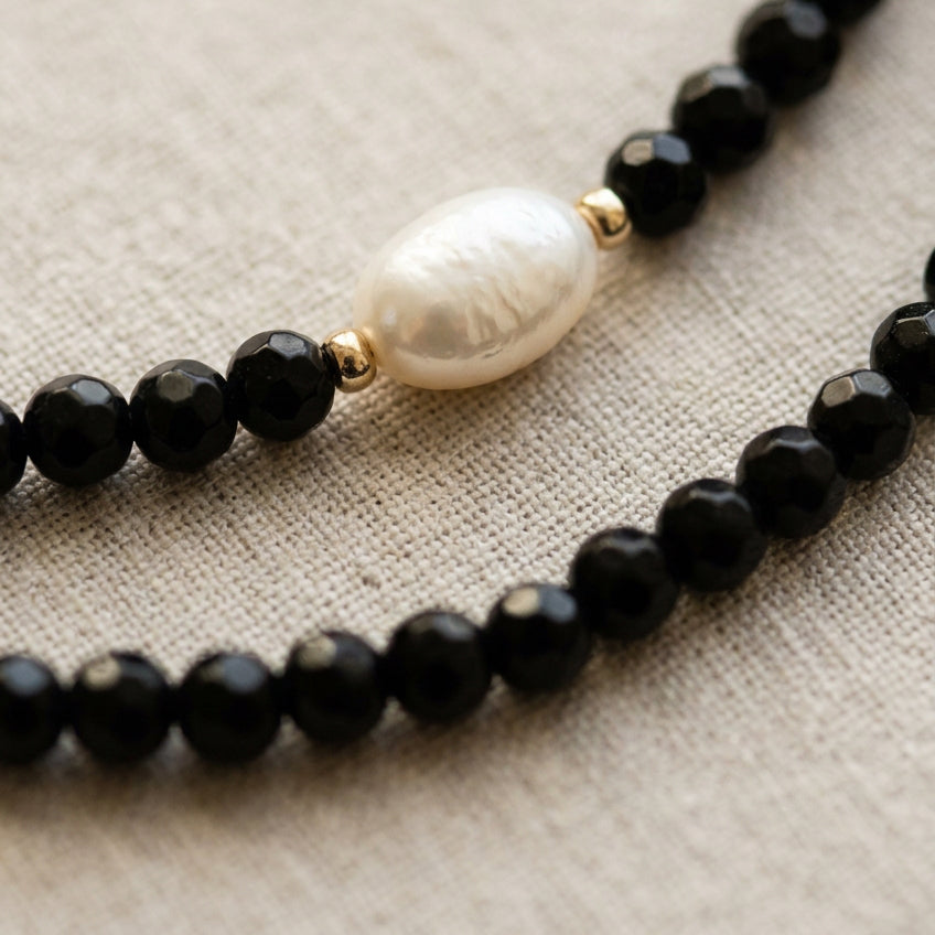 Close-up of a white rice-shaped baroque pearl between gold-coloured spacers and faceted black onyx beads, with a second black onyx strand resting alongside on cream linen.